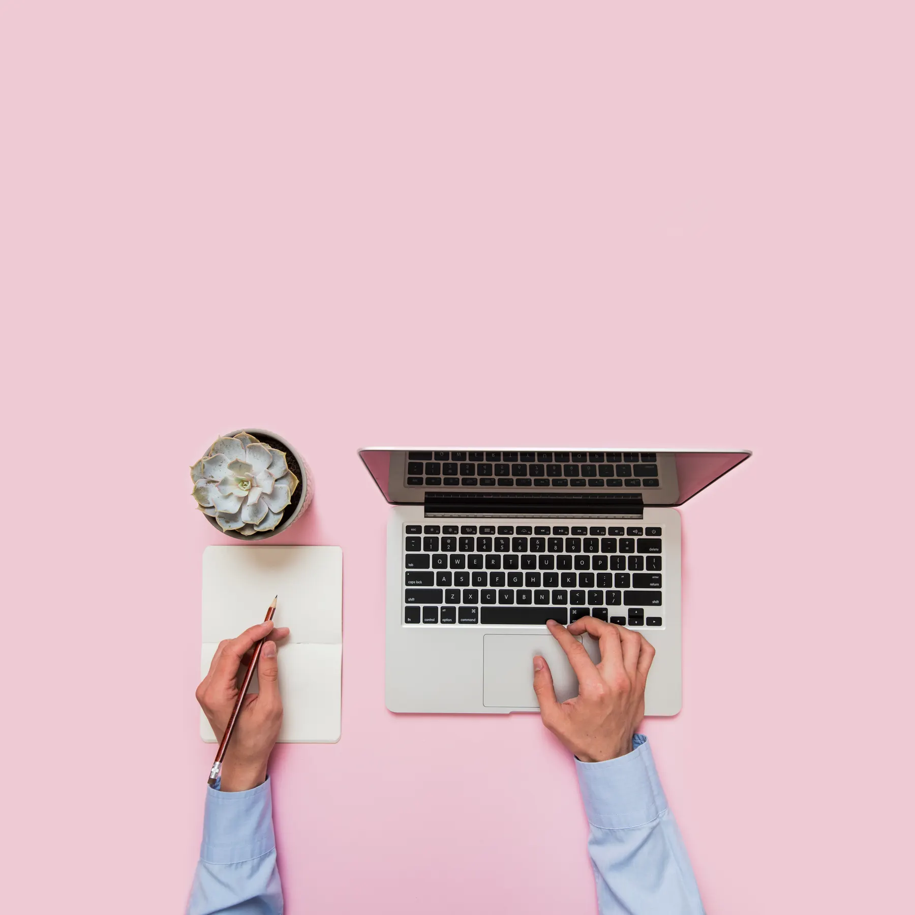 Manos escribiendo en una laptop y una libreta sobre un fondo rosa