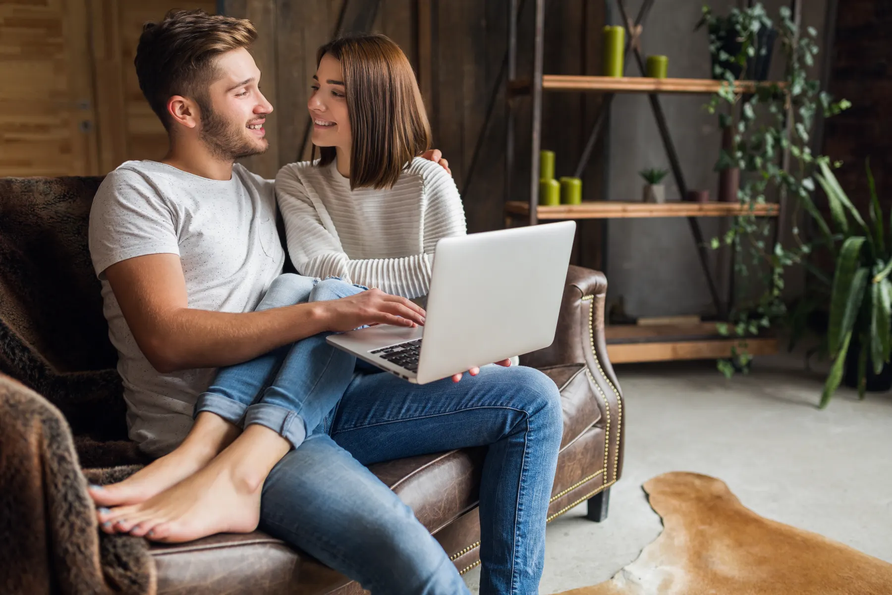 Pareja sonriendo mientras comparte una laptop en el sofá de casa